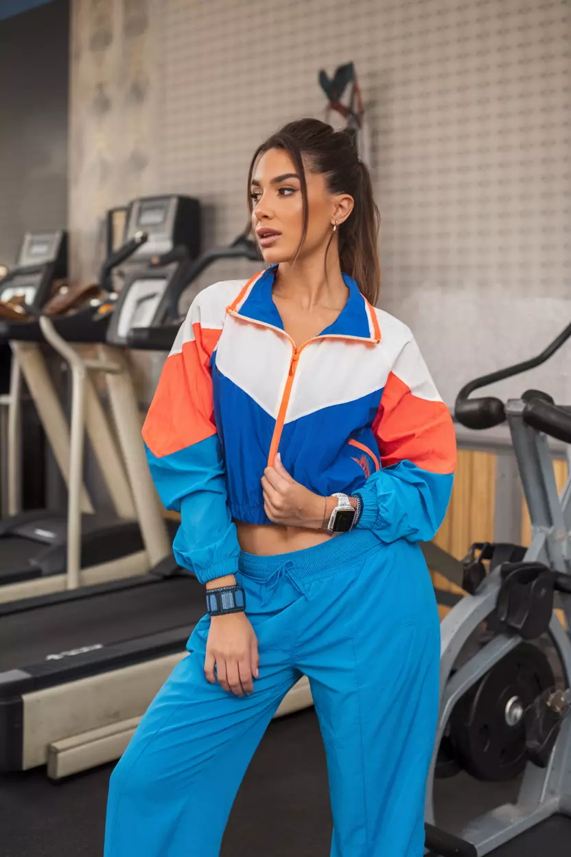A beautiful woman wearing a stylish colorful windbreaker with bright blue track pants, she wears a digital watch and sweatbands, retro gym setting